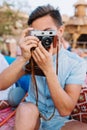 Portrait of boy with short black hair, holding retro camera and making photo, while sitting in open-air cafe. Young man Royalty Free Stock Photo