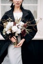 Portrait of a bride in a white silk wedding dress and a black coat with a bride`s bouquet in her hands. Black wooden Royalty Free Stock Photo