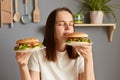 Portrait of brown haired Caucasian woman holding two delicious fresh hamburgers, smelling aromat of fast food with closed eyes and Royalty Free Stock Photo