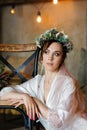 Portrait of a brunette bride with a wedding hairstyle and a flower wreath, in a white transparent negligee. Morning of the bride