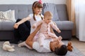 Portrait of brunette father playing with his daughters while lying on floor near sofa, wearing white t shirt, children sitting on Royalty Free Stock Photo