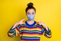 Portrait of brunette girl pointing blue mask from corona wear rainbow sweater isolated on bright yellow color background