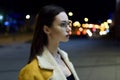 Portrait of brunette in glasses with face turned to the side and lit by city centre lights. Womenswear fashion