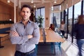 Portrait Of Businessman Standing In Busy Modern Open Plan Office With Colleagues In Background