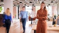 Portrait Of Businesswoman By Desk In Busy Multi-Cultural Office With Motion Blurred Colleagues