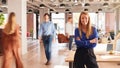 Portrait Of Businesswoman By Desk In Busy Multi-Cultural Office With Motion Blurred Colleagues