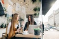 Portrait through cafe window, two happy business women sitting in cozy cafe with laptop talking over smiling. Two young ladies in