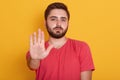 Portrait of calm bearded young man wearing red casual t shirt standing with stop warning gesture hand and looking at camera with