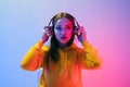 Portrait of a calm Caucasian woman wearing a yellow hoodie sitting and wearing wireless headphones, and posing isolated on a neon