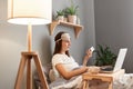 Portrait of calm serious woman lying in bed, holding mobile phone in hands, working on laptop computer, typing on keyboard, Royalty Free Stock Photo