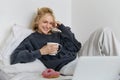 Portrait of candid, happy young woman lying in bed, looking at laptop screen, holding cup of tea and eating doughnut Royalty Free Stock Photo