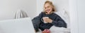 Portrait of candid, happy young woman lying in bed, looking at laptop screen, holding cup of tea and eating doughnut Royalty Free Stock Photo