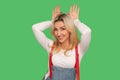 Portrait of carefree beautiful adult blond woman in stylish denim overalls showing bunny ears and smiling