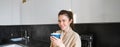 Portrait of carefree smiling woman drinking coffee, standing in kitchen with delighted, pleased face expression, waking