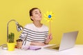 Woman blowing at windmill while sitting on workplace with laptop, playing with pinwheel toy on stick Royalty Free Stock Photo