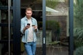 Portrait of casual relaxed man using phone for online payment on doorstep of his home, shopping online from home leaning on door