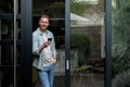 Portrait of casual relaxed man using phone for online payment on doorstep of his home, shopping online from home leaning on door