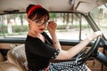 Portrait of Caucasian beautiful young girl in black vintage dress and vintage glasses posing behind the wheel of a vintage car Royalty Free Stock Photo