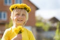 Portrait of Caucasian blond boy with wreath of dandelions and bouquet of flowers near the house. Blurred background Royalty Free Stock Photo