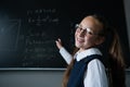 Portrait of a caucasian girl in glasses in the classroom. The schoolgirl writes the formula with chalk on the blackboard