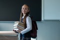 Portrait of a caucasian schoolgirl with a backpack. The girl is holding a stack of textbooks in the classroom.