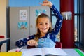 Portrait of caucasian schoolgirl sitting at desk raising hand in classroom at elementary school Royalty Free Stock Photo