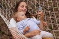 Portrait of Caucasian woman with her infant kid in forest, mother and daughter lying in hammock, female using mobile phone, Royalty Free Stock Photo