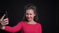 Portrait of caucasian woman in red smilingly making selfie-photos using smartphone on black background.