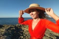 Portrait of a Caucasian young woman in a red dress and straw hat on the seashore. holds a hat with his hands and smiles Royalty Free Stock Photo