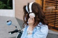 Portrait of charming girl with shiny curly dark-brown hair enjoying favorite music in big white earphones on moped. Shy Royalty Free Stock Photo