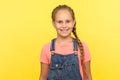 Portrait of charming little girl with long braid in denim overalls looking at camera with toothy smile Royalty Free Stock Photo