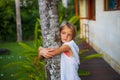 Portrait. Charming little girl standing and holding the tree, wearing white dress. Cute face. Spending time outdoor in Royalty Free Stock Photo