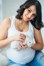 Portrait of the charming pregnant woman with beautiful smile and dark curly hair petting little white fluffy rabbit.