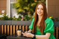 Portrait of charming redhead young woman holding mobile phone sitting at table with coffee cup in outdoor cafe terrace Royalty Free Stock Photo