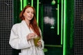 Portrait of charming redhead young woman in white bathrobe, holding glass with freshly squeezed vitamin juice in hands Royalty Free Stock Photo