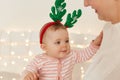 Portrait of charming toddler baby girl wearing striped sleeper and deer hoop, sitting in mother arms, touching her mommy, posing