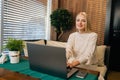 Portrait of charming young woman with long blonde hair sitting at table in cafe while using laptop computer and smiling Royalty Free Stock Photo