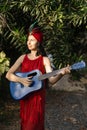 Portrait of young woman in red dress, turban with feathers with blue guitar in her hands in park. Royalty Free Stock Photo