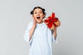 Portrait of cheerful amazed young dark-haired girl holding in hand gift having fun, standing in trendy t shirt over light grey