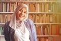 Portrait of cheerful Asian muslim female librarian wearing hijab and smiling, woman standing against books in library