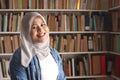 Portrait of cheerful Asian muslim female librarian wearing hijab and smiling, woman standing against books in library