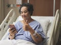 Cheerful  Asian senior  female patient  sitting  in hospital bed,  smiling and looking at mobile phone, video calling  and showing Royalty Free Stock Photo