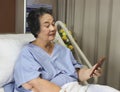 cheerful Asian senior female patient sitting in hospital bed, smiling and looking at mobile phone, video calling to her Royalty Free Stock Photo