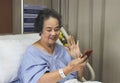 Cheerful  Asian senior  female patient  sitting  in hospital bed,  smiling and looking at mobile phone, video calling  and waving Royalty Free Stock Photo