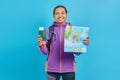 Portrait of cheerful asian young man wearing purple jacket showing map and passport book isolated on blue background