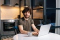 Portrait of cheerful bearded young man using typing on laptop sitting at table in kitchen room with modern interior Royalty Free Stock Photo