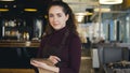 Portrait of cheerful and beautiful young waitress standing, keeping menu, and smiling sincerely in slylish cafe.