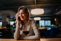 Portrait of cheerful blonde female talking on mobile phone sitting at table with red wine in modern restaurant with dark Royalty Free Stock Photo