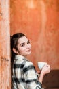 Portrait of a cheerful brunette girl holding a cup of coffee while leaning on a wall.