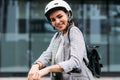 Portrait of cheerful businesswoman in white cycling helmet looking at camera leaning on handlebar of electric push scooter